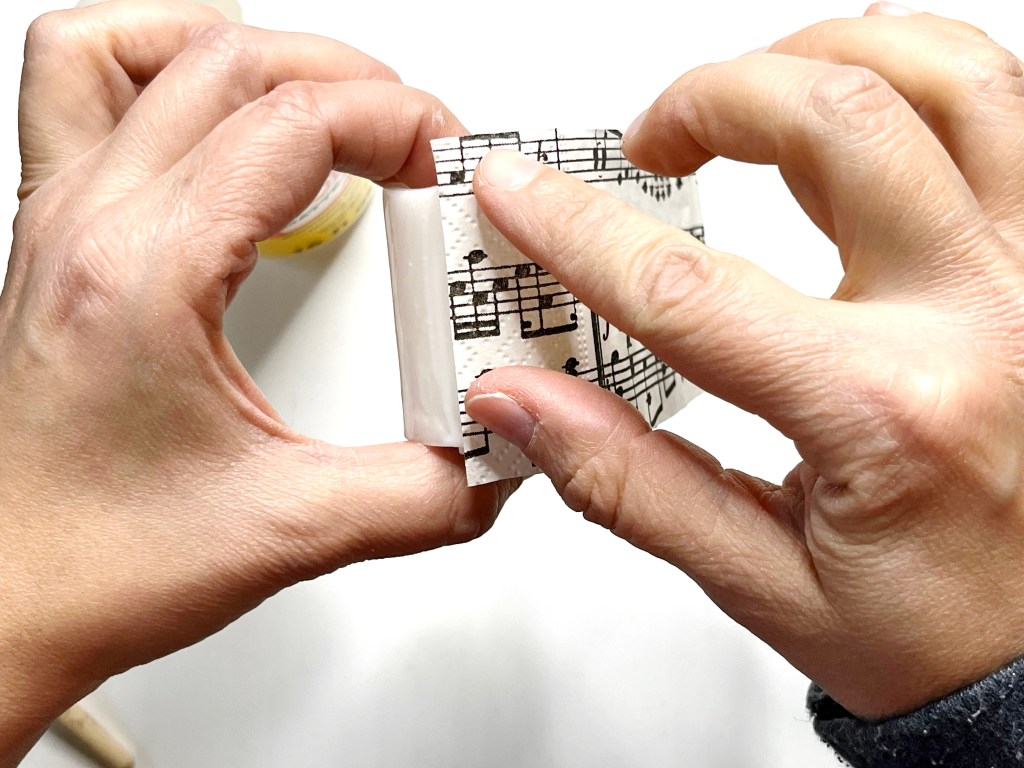 Music themed napkin rectangles is placed on the battery operated candle covered in glue.