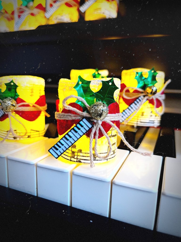 Several music note candles sitting on a piano keyboard. The candles are lit up. The candles have red ribbon and twine tied around them and have a keyboard charm, holly leaves and a jingle bell as embellishments.
