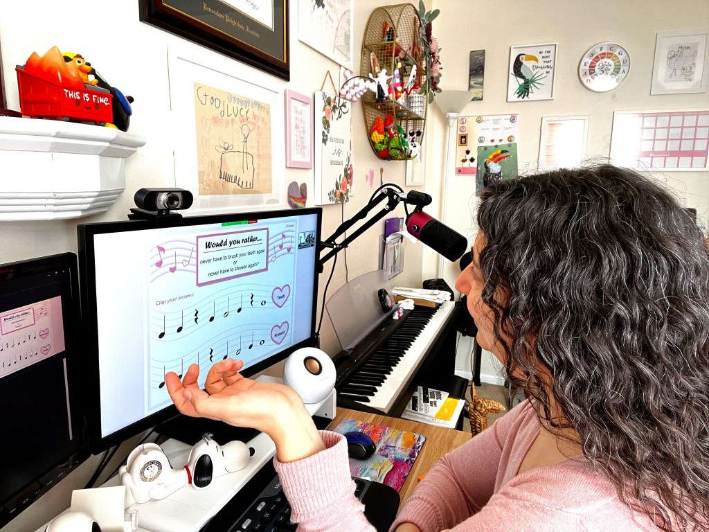 Piano teacher in a pink sweater teaching a student during an online lesson. They are playing a Valentine's day rhythm game that show quarter notes, quarter rests, half notes, and whole notes.