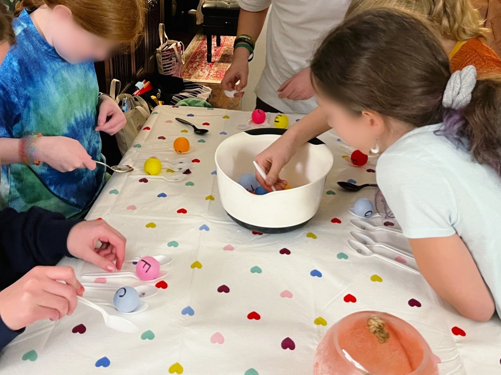 Children gathered around a ball filled with ping pong balls. They are playing Best in Pass a note-reading race game. One child is attempting to scoop a ping pong ball out of the bowl with a white plastic spoon. Two other children are trying to move a ping pong ball from one spoon to another.