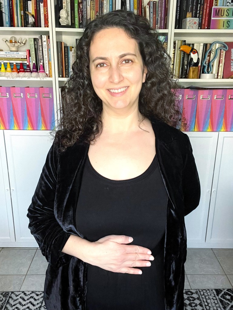 Woman with curly brown hair, dressed in a black jumpsuit and blazer with her hand on her stomach to show proper hand placement when bowing. Front view.