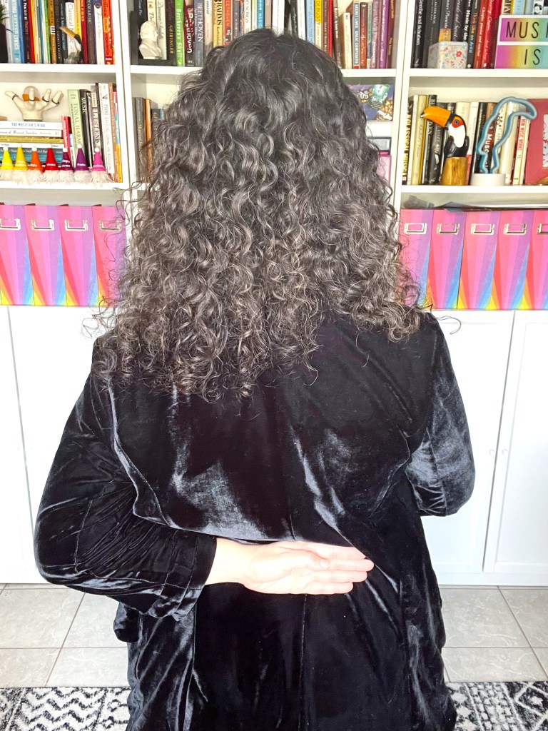 Woman with curly brown hair, dressed in a black jumpsuit and blazer with her hand on her back to show proper hand placement when bowing. Back view.