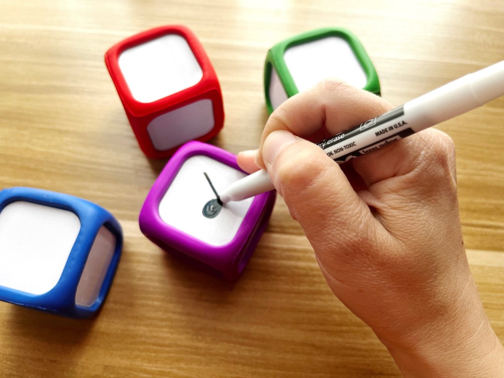 A hand holds a black dry-erase marker. It is drawing a quarter note on a purple dry-erase die. Three other dice sit in the background, a blue one, a red one, and a green one.