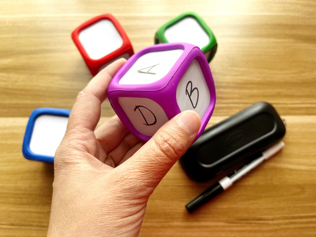 Hand holding a purple dry erase die with the letters A, B and D written on its sides in black marker. In the background are three other dice (a red one, a blue one and a green one) along with a dry-erase marker and eraser.