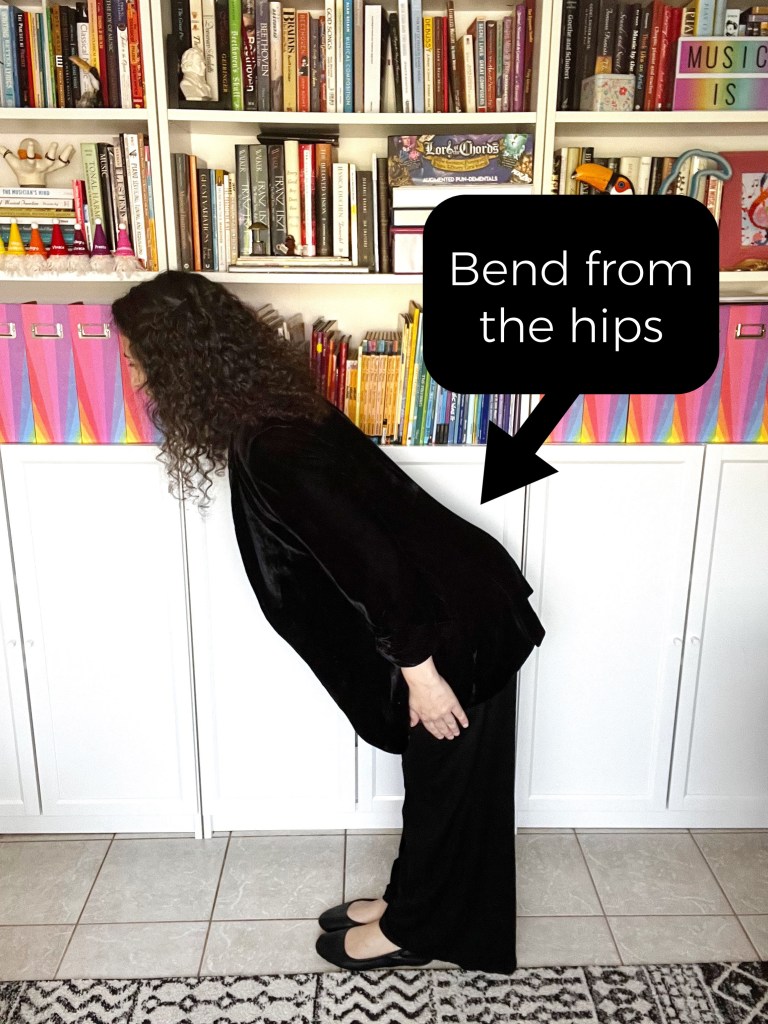 Woman with curly brown hair, dressed in black pants and blazer, bending at the hips to show the proper body position for bowing.