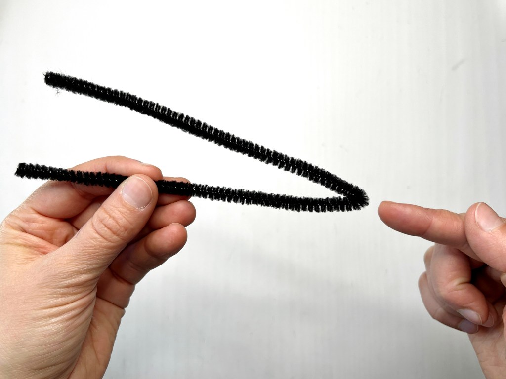 A black pipe cleaner has been bent in half and a hand points to the bend.