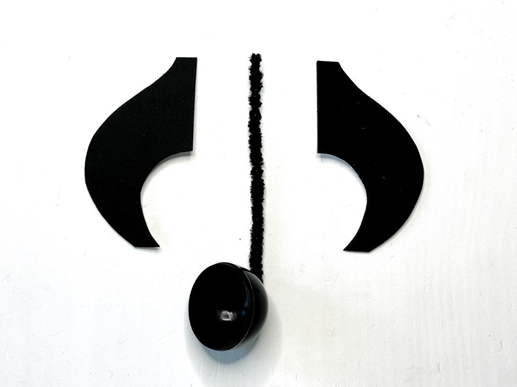 Half of a black plastic egg with a black pipe cleaner attached to it sit between two black eighth note flags made of self-adhesive craft foam