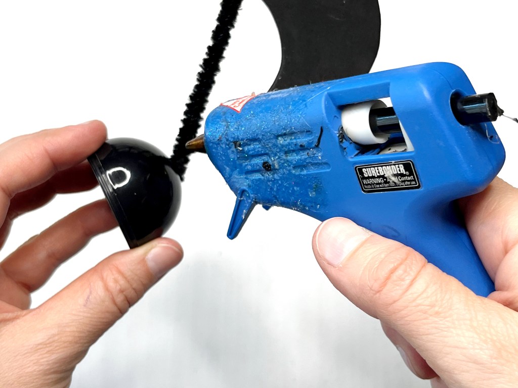 A hot glue gun loaded with a black hot glue stick applies glue to the base of the plastic egg music note