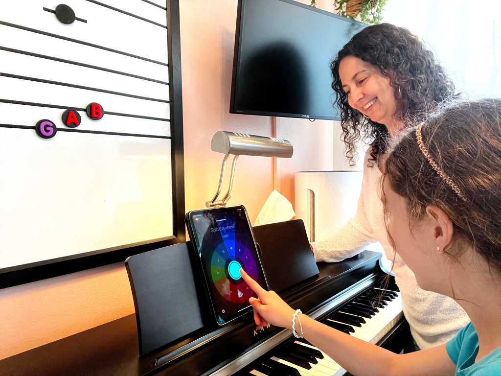 Piano teacher stands next to a piano as a student prepares to tap the screen of a tablet with a rainbow colored wheel of fortune on it.