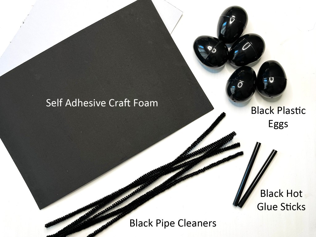 Black self adhesive craft foam sheets, five black plastic eggs, a bunch of black pipe cleaners, and two stick of black hot glue sit on a white table.