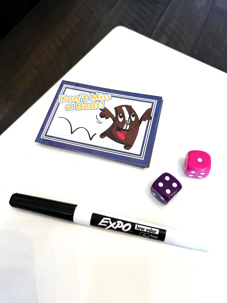 Don't Miss a Beat! rhythm card deck sits on top of a whiteboard and next to a pair of dice and a black dry-erase marker.