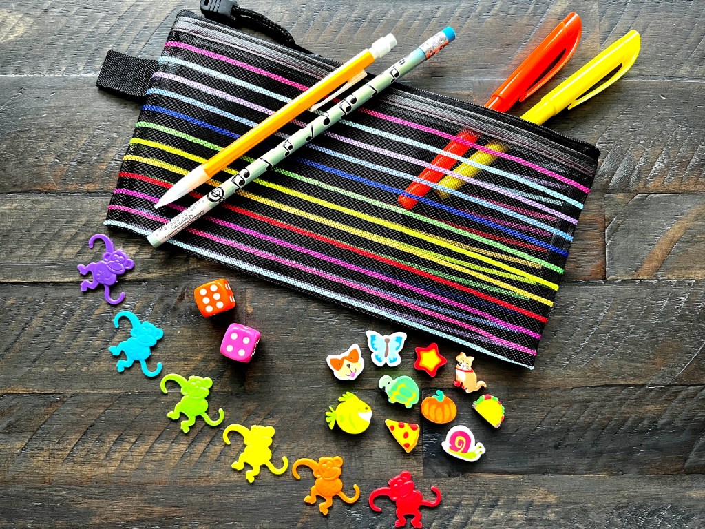 Black rainbow piano practice pouch lays on a wood table with its contents spread out around it: orange highlighter, yellow highlighter, mechanical pencil, music pencil, two dice, rainbow barrel monkey, mini-erasers.