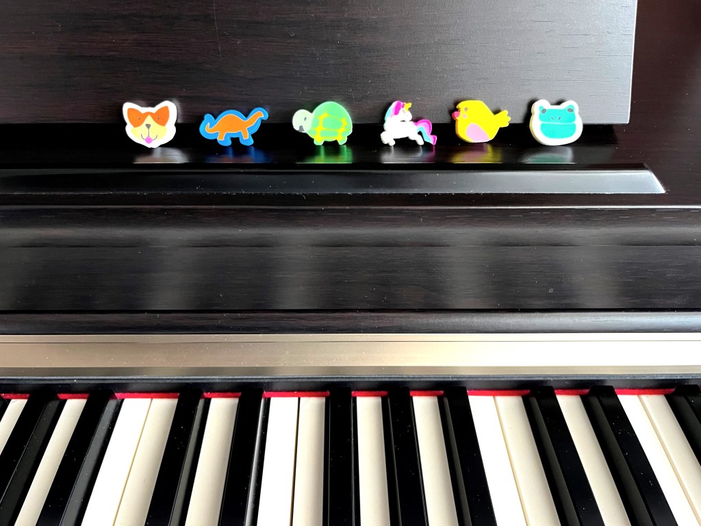 6 animal mini-erasers are lined up along the book rack of a piano.