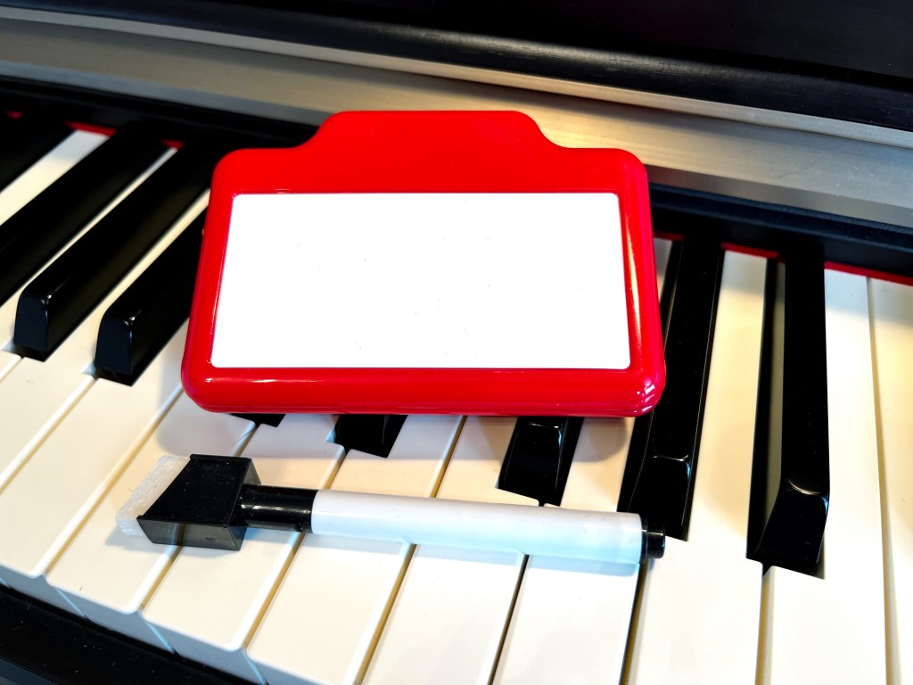 A red dry erase magnetic clip sits on top of the keys of a piano. A dry erase marker with an eraser sits next to the clip.
