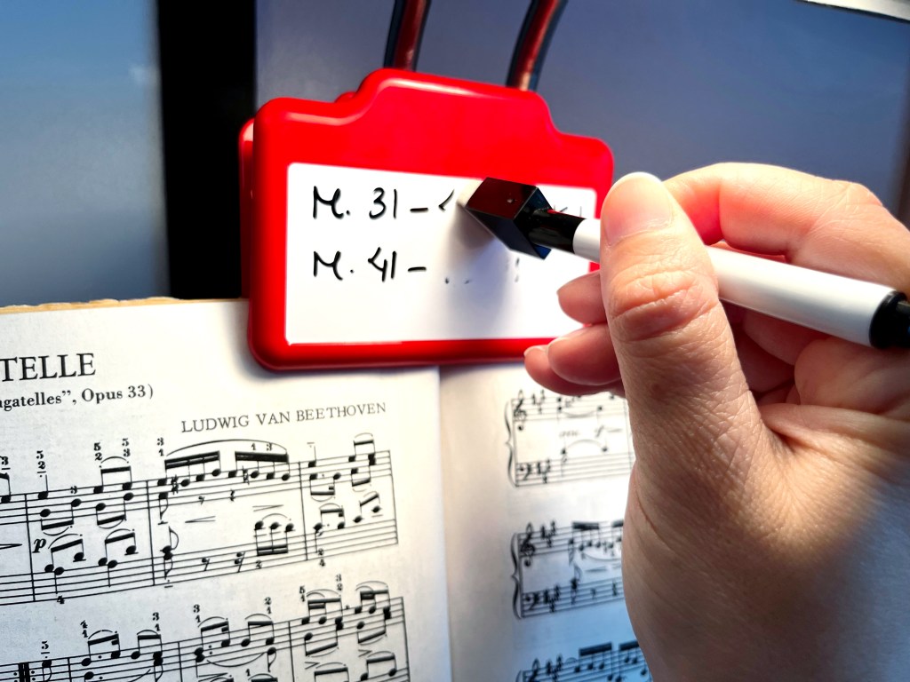A hand is holding a dry erase marker and erasing the writing on a dry erase board clip that is clipped to a book of music by Beethoven.