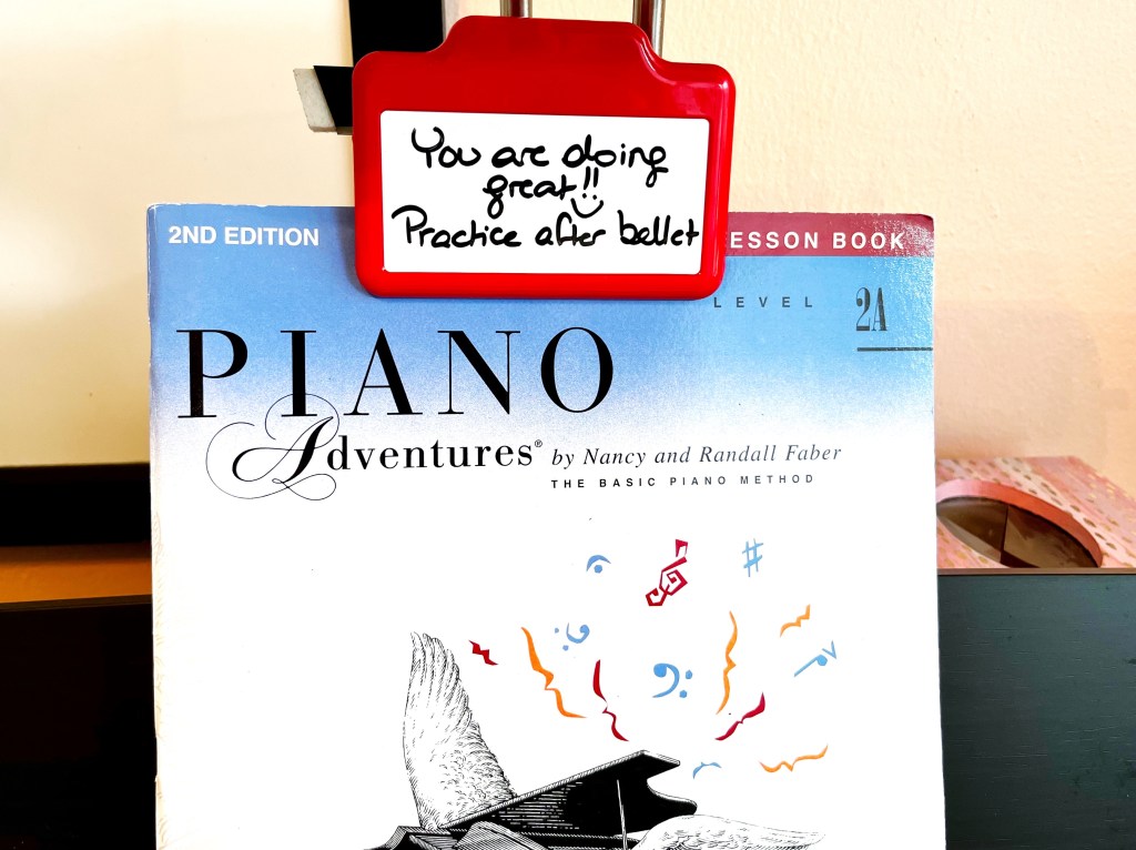 A dry erase clip is clipped to the top of a piano book. It has a message on it that reads, "You are doing great!! Practice after ballet".