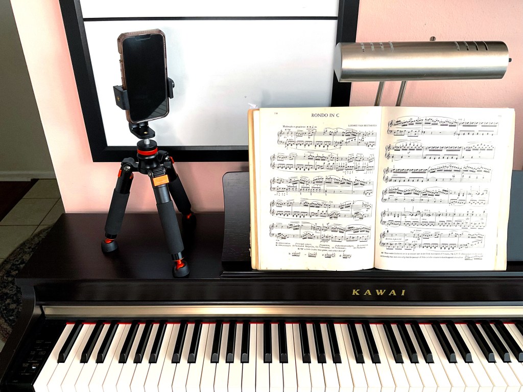 A tabletop tripod holding a smartphone sits on top of a piano