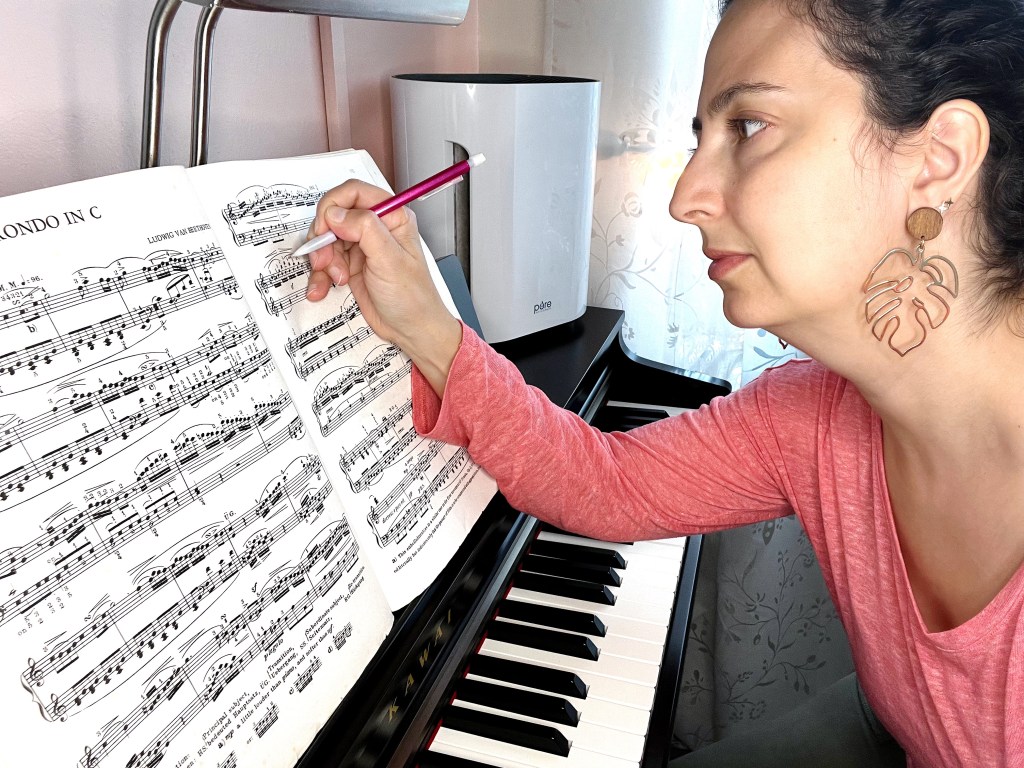 Dark haired woman in a pink sweater sitting at a piano makes notes in her music score