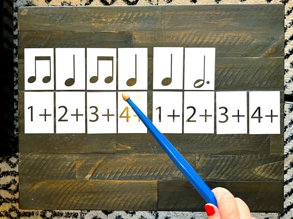 Rhythm cards sit on a dark brown wooden table as a piano student hold a blue drumstick in her hand to tap out the rhythms.