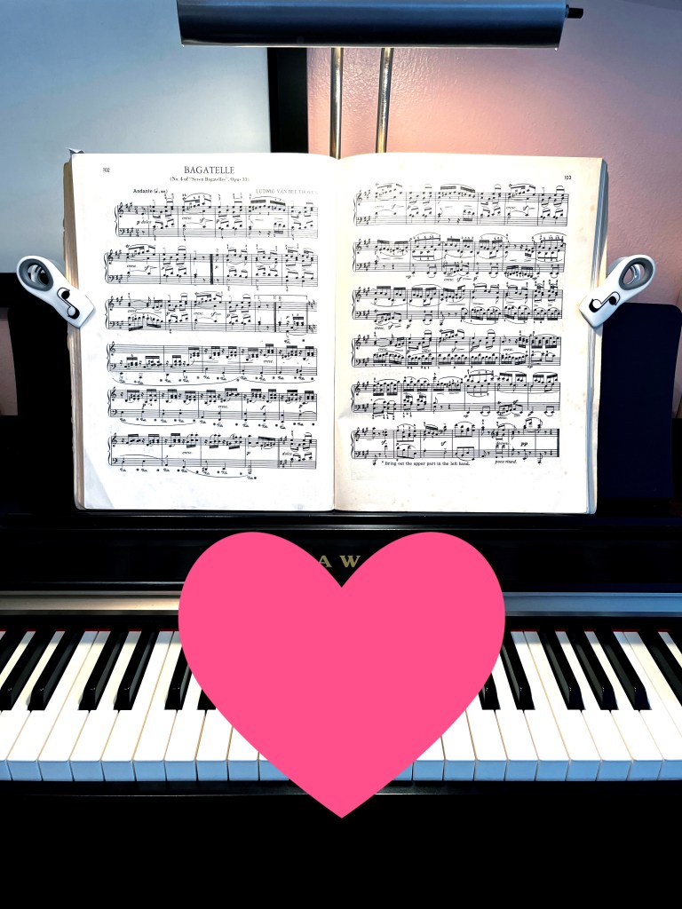 A piano book on the book rack of a piano. Music clips hold the book open by holding it against the book rack. A large pink heart indicates this set up works for keeping music in place.