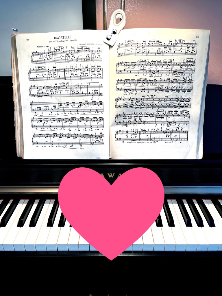 A piano book on the book rack of a piano. Music clips hold the book open by gripping the spine of the book. A large pink heart indicates this set up works for keeping music in place.