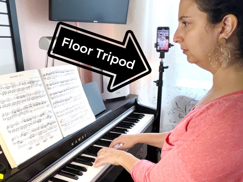 Dark haired woman in a pink sweater play the piano next to a floor tripod that is recording her playing