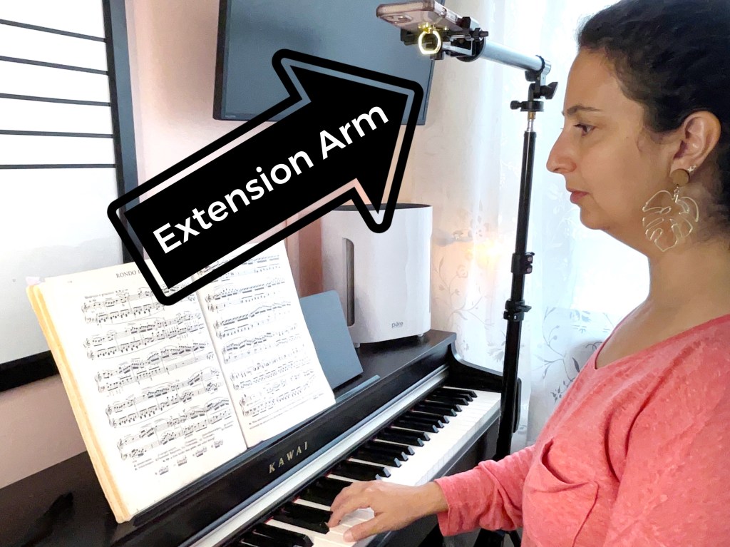 Dark haired woman in a pink sweater play the piano next to a floor tripod with an extension arm that is recording her playing