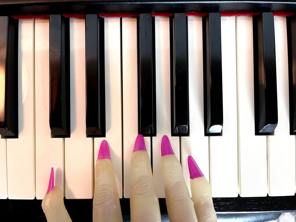 A zombie hand with bright pink fingers rests on the keys of a piano.