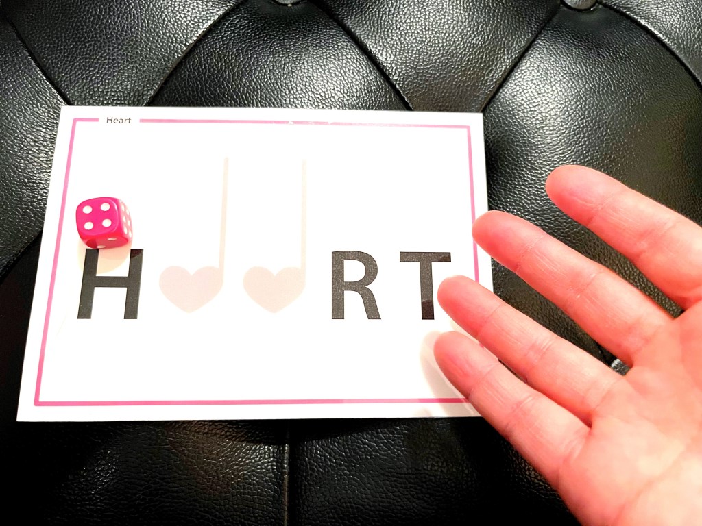 A piano student rolls a pink die on top of a card that reads "Heart" but has letters missing. 