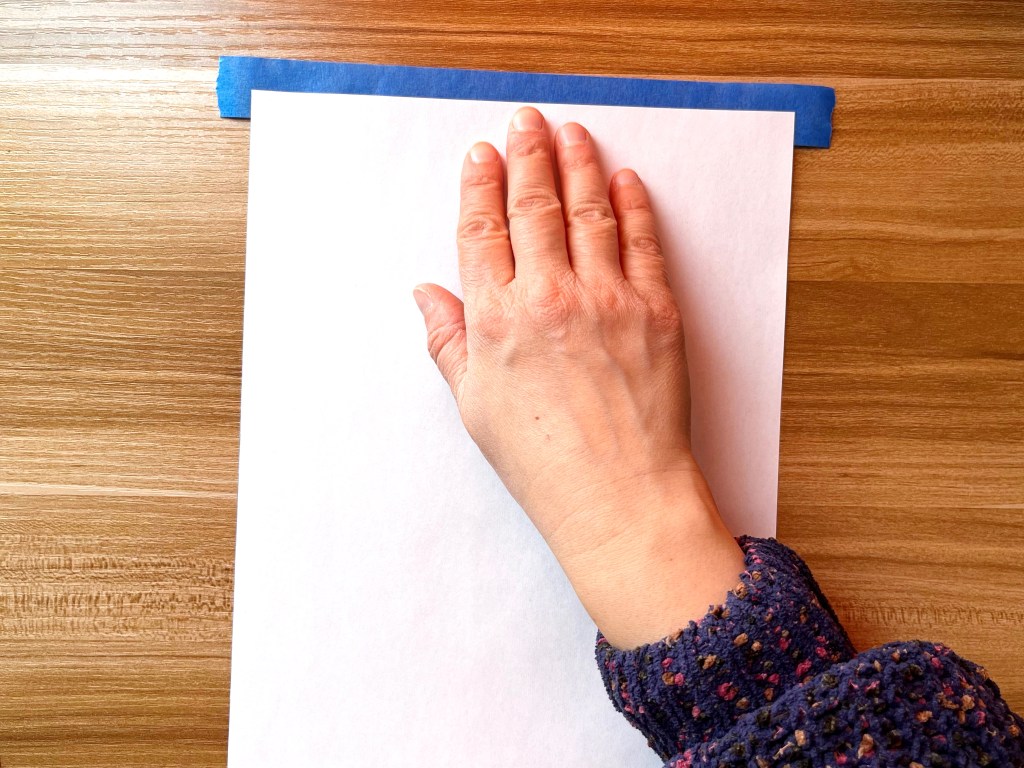 A person tapes together a sheet of copier paper and a sheet of white tissue paper using blue painter's tape.