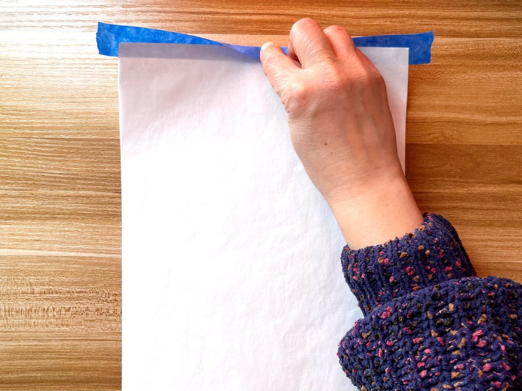 A person folds over a strip of blue painters tape to secure two sheets of paper together.