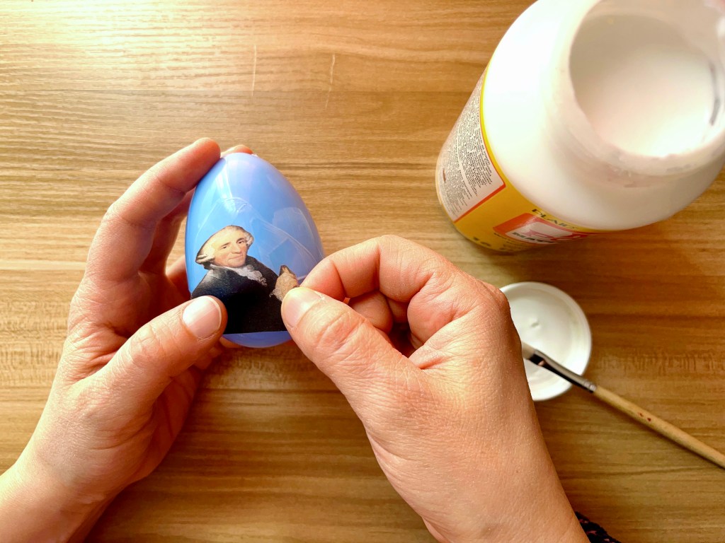 A person glues an image of Joseph Haydn to a blue plastic easter egg.