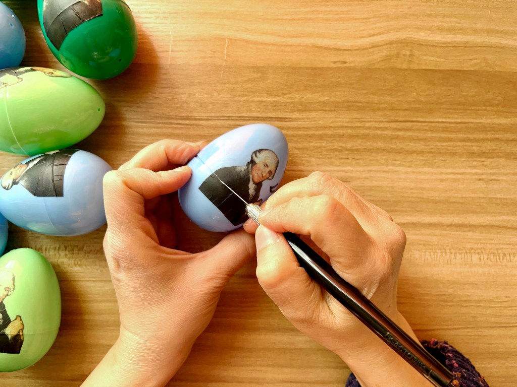 A person cuts the image of Joseph Haydn along the seam of a blue plastic easter egg using an xacto utility knife.