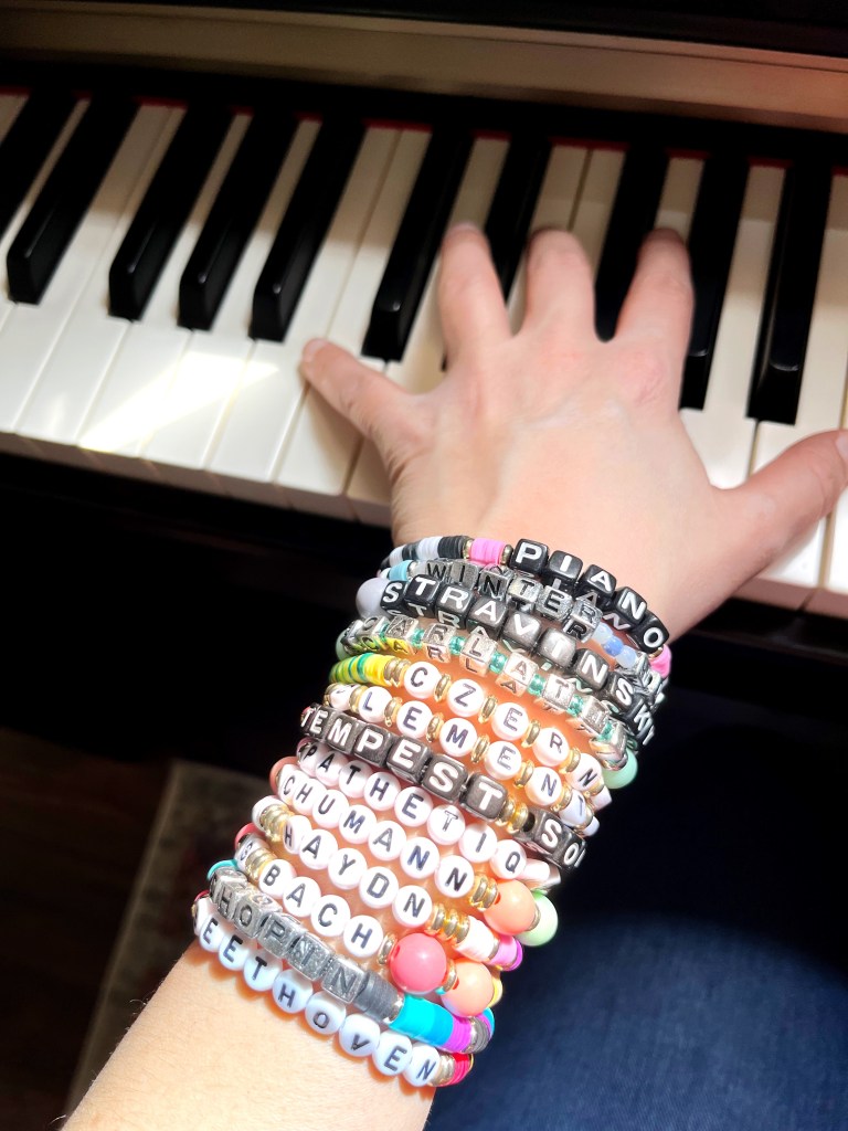 A hand sits on the keys of a piano. The arm is covered with a stack of colorful friendship bracelets with the names of composers and compositions on them.