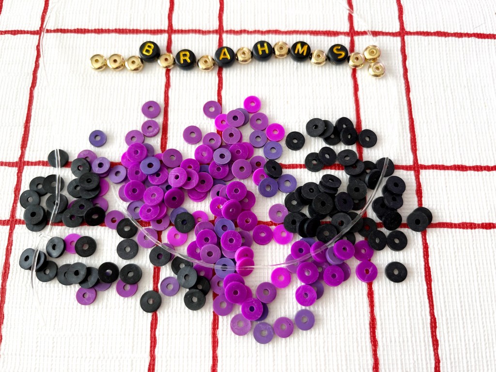 Purple, gold and black beads lay on a table. Letter beads spell out the name Brahms.