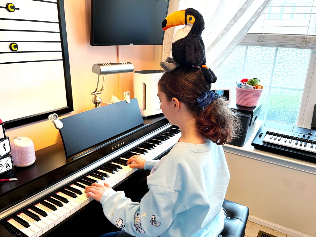 Piano student plays the piano while balancing a stuffed toucan on her head.