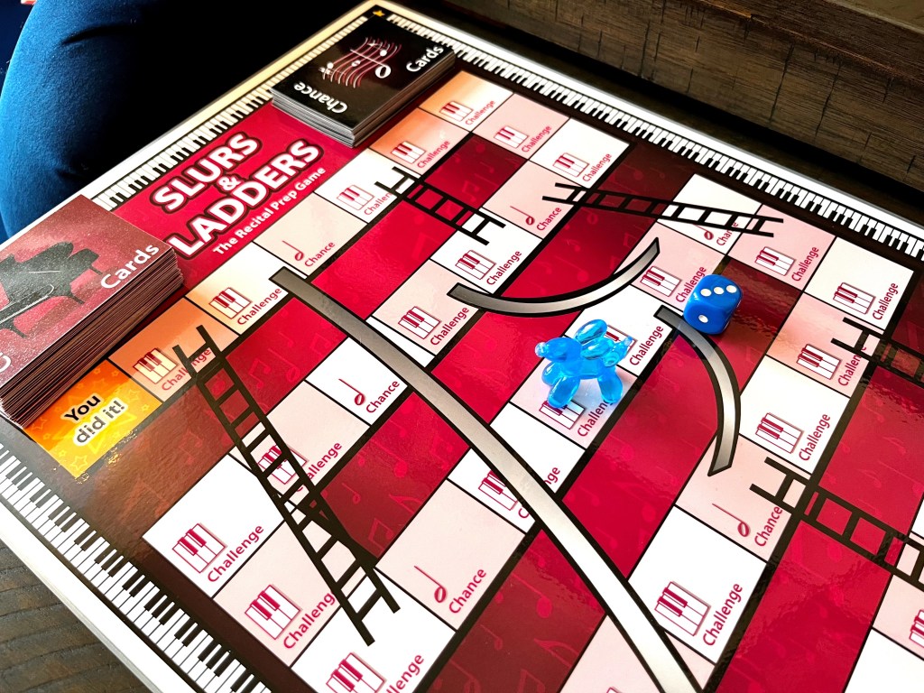 Slurs and Ladders, the piano recital prep game, sits on a wood table along with a blue balloon dog game marker and a blue die.
