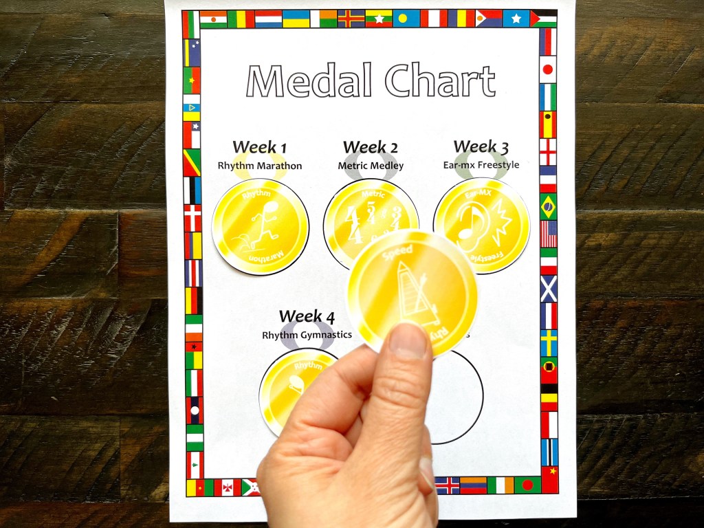 A hand hold a gold medal in the Rhythm Olympics over a medal chart filled with other gold medals.