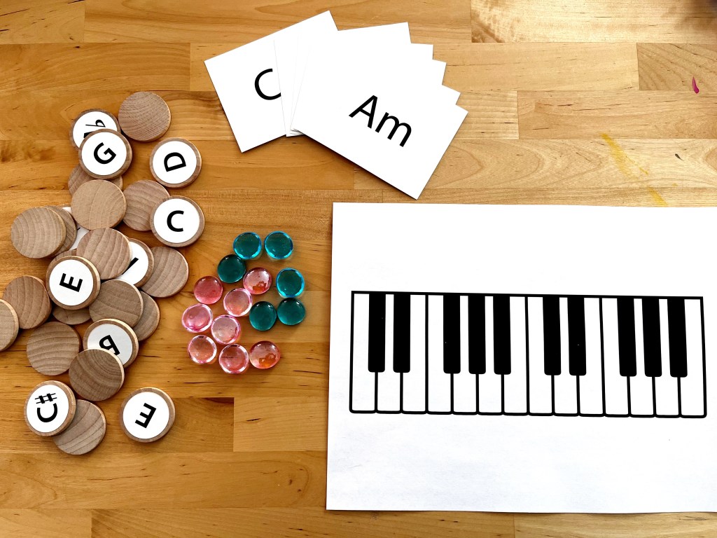 Supplies for a chord games are laid out on a table. The supplies include chord cards, a keyboard printout, wooden circles with pitches printed on them, and blue and pink gems.