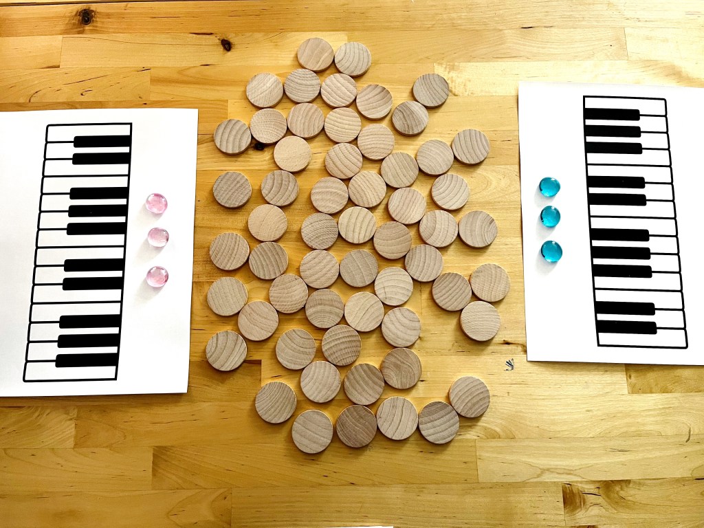 Three Chord Dash, a chord game, is set up on a table. Each team has a keyboard printout with 3 gems lined up along the top of the printout. Wooden circles are arranged at the center of the table between the two printables.