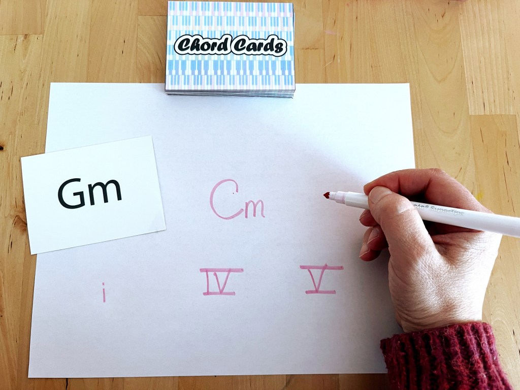 A piano student writes out the four and five chords in the key of G minor. A deck of chord cards sits on the table above the paper.