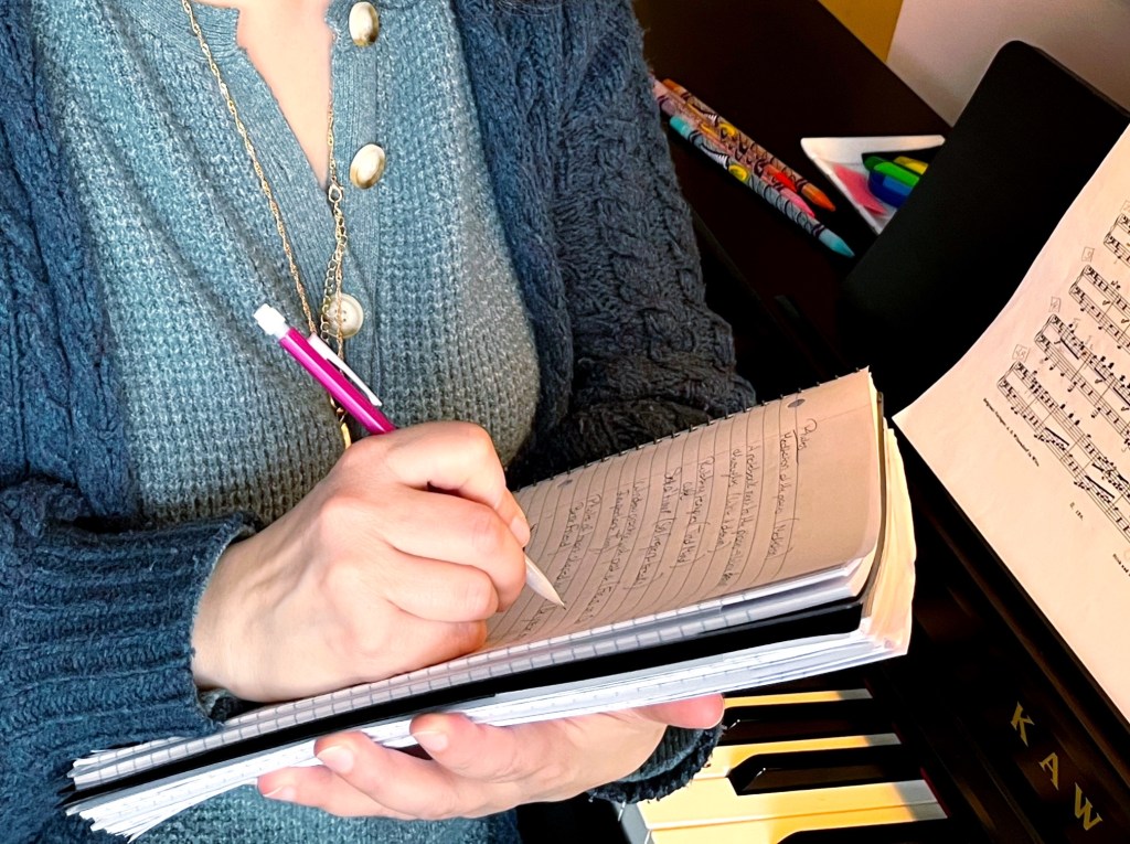 A piano students writes down their thoughts in a notebook with a pink pencil while sitting at the piano.