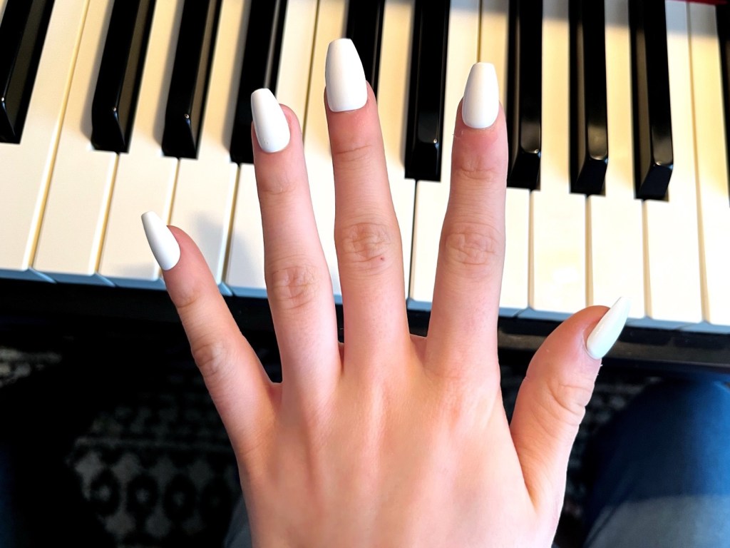 A hand with white fake press on nails is outstretched over the keys of a piano