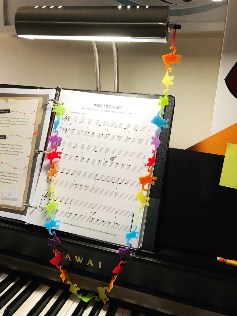 A colorful chain of barrel monkeys hangs from the ring of a binder and a piano lamp, above some sheet music.