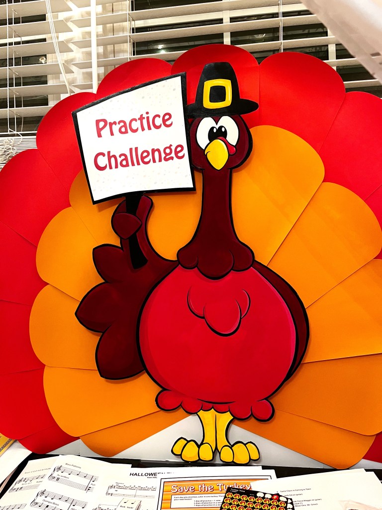 A colorful turkey decoration holding a sign that says 'Practice Challenge', set against a backdrop of layered orange and red feathers.